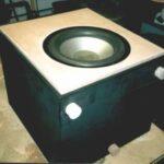 Ein auf dem Rücken liegender Subwoofer in schwarzer Lackierung, dem eine Holzfront angepast wird.