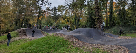 Eine Pumptrackanlage in einem Park. Auf der Anlage fahren etliche Jugendliche mit Fahrrädern und Scootern. Viele stehen in einer Schlange und warten, bis sie an der Reihe sind.