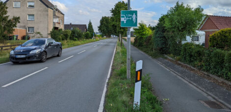 Eine zweispurige Straße mit einem einseitig angelegten Gehweg. Ein Zusatzschild an einem Laternenmasten weist auf den laut StVO vorgeschriebenen Mindestabstand von 1,50 Metern beim Überholen von Fahrrädern hin.