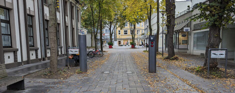 Heruntergefahrene Poller am Eingang einer Fußgängerzone. Man sieht eine von Bäumen rechts und links gesäumte Straße mit durchgängiger Bebauung dahinter.