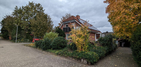 Ein Haus hinter Büschen versteckt. Der Himmel ist wolkig, Bäume rechts und links im Bild haben herbstlich buntes Laub.