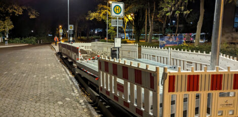 Ein Bushaltestellenschild auf einem Omnibusbahnhof, welches goßflächig mit Absperrschranken umzäunt ist, weil ringsherum Baumaterial liegt.