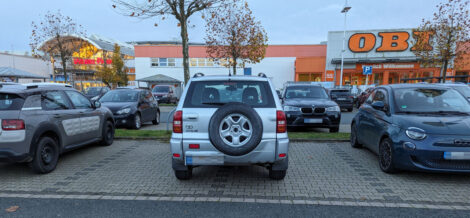Ein SUV parkt auf einem Baumarktparkplatz genau mittig auf der Linie zwischen zwei gekennzeichneten Parkflächen. Rechts und links stehen Fahrzeuge korrekt in den Parkbuchten.