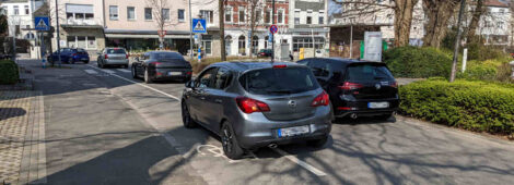 Eine Autoschlange vor einer Einmündung einer Straße. Auf einem links markierten Fahrradstreifen stehen ebenfalls Autos.