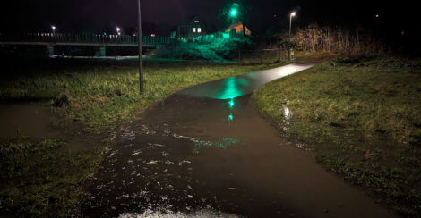 Ein auf einer Strecke von einigen Metern unter Wasser stehender Radweg durch eine in einer Mulde gelegenen Wiese.