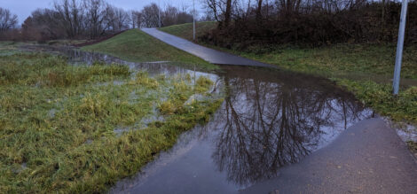 Eine große Wasserfläche auf einem asphaltierten Geh- und Radweg vor einer Böschung, auf welcher Weg per Rampe aufwärts führt.