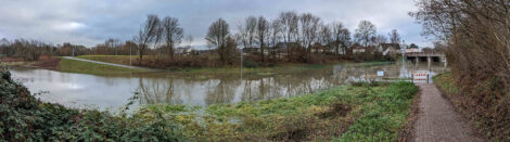 Ein über die Ufer getretener Fluss, der die Ausdehnungsfläche einer Flutmulde nutzt. Dabei wird ein Geh- und Radweg unter Wasser gesetzt. Auf der anderen Seite sieht man die Rampe des Weges im Wasser enden.