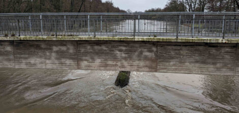 Eine Brücke über einem Fluß. Man schaut auf die Wasserfläche und sieht eine Stütz der Brücke, die gerade noch aus dem Wasser schaut.