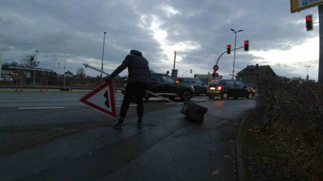 Ein Mann hebt einen Schilderpfahl und das dazugehörige, aber abgefallene Schild "Bodenwellen" hoch, weil es quer über einen Geh- und Radweg liegt.