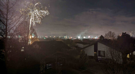 Feuerwerk an Silvester von einem Balkon aus gesehen. Man sieht am Horizont viele Raketen aufsteigen. Im linken vorderen Teil des Bildes explodiert gerade eine deutlich nähere Rakete.