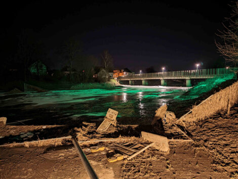 Eine grün beleuchtete Eisfläche auf einer überfluteten Wiese wird im Hintergrund von einer Brücke begrenzt. Vorne sieht man umgeworfene und von Schlamm bedeckte Schilderpfähle und Bauzäune.