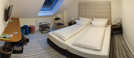 Ein hell eingerichtetes Hotelzimmer mit einem Doppelbett, einem kleinen Beistelltisch neben einem Sessel und einem Board an der Wand.