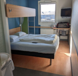 Ein kleines Hotelzimmer mit einem Doppelbett und einem quer über dem Kopfende montierten Querbett. Links im Vordergrund ist das im Zimmer befindliche Waschbecken zu sehen.