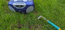Ein kleines Kofferradio auf einem grünen Rasen aus dem zwei Ende eines Leitdrahtes für Rasenmäherroboter heraus schauen. Daneben sieht man ein Werkzeug, um Rasenkanten abzustechen.