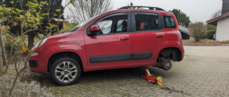 Ein roter Fiat Panda ist mit einem Wagenheber hinten angehoben und das linke Hinterrad ist entfernt.