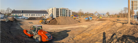 Auf einer großen Freifläche sind Planierraupen und Bagger unterwegs und verschieben Erde. Im Hintergrund sieht man ein Parkhaus neben einem langezogenen, etwas niedirgeren Haus.