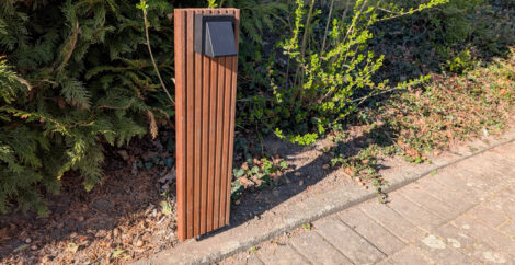 Vor einer Hecke steht senkrecht ein kleines Brettchen aus Bangkiraiholz. Am oberen Ende des Brettes ist ein kleiner, schwarzer LED-Strahler befestigt.