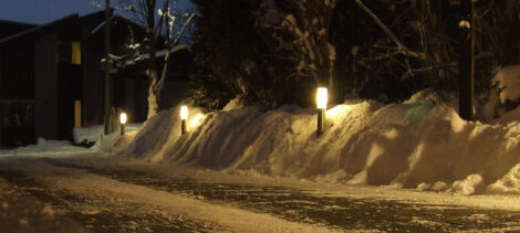 Aus einem Schneewall entlang der Zufahrt zu einem Haus schauen Gartenleuchten heraus und erhellen den Schnee und die weiße Einfahrt in warmweißem Licht.