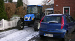 Ein blauer "New Holland"-Traktor steht in einer Hofeinfahrt, welche von einem weißen Lattenzaun begrenzt wird. Auf einem Stellplatz daneben steht ein blauer Fiat Punto.