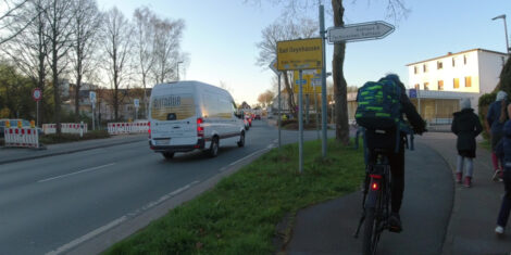 Auf einem Radweg fährt ein Schüler neben zu Fuß Gehenden auf dem Gehwegteil vorbei. Der Weg ist nicht besonders breit. Rechts ist eine Hecke, links ein Grasstreifen zur Fahrbahn hin.