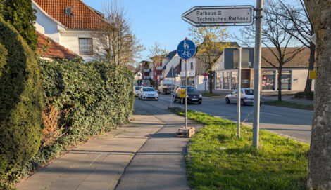 Auf einem viel zu schmalen Radweg steht hinter einem Knick ein provisorisches Verkehrszeichen 240, um anzuzeigen, dass der viel zu schmale Radweg auch in Gegenrichtung benutzungspflichtig ist