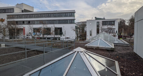 Blick über die obere Etage eines Einkaufszentrums mit pyramidenförmigen Lichtschächten im Dach, welches mit Kies bedeckt ist. Im Hintergrund ist die Fassade eines großen Gebäudes mit vielen Fensterflächen zu sehen.