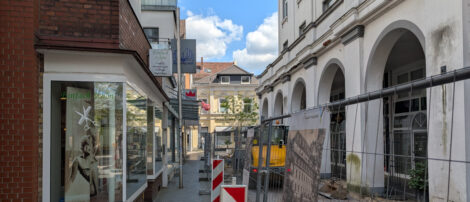 Eine sehr schmale Fußgängerzone, mit einer Kolonade rechts und Geschäften inks. Fast die gesamte Verkehrsfläche ist mit einem Bauzaun abgesperrt. Mitten im verbleibenden Bereich für zu Fuß Gehende, steht ein mobiler Fahnemast, an dem nichts weiter befestigt ist.