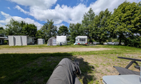 Auf einem Campingplatz stehen vor einer Reihe Bäume und Büsche ein paar Caravans. Im Vordergrund sieht man die übereinandergeschlagenen Beine des Fotografen - mir!