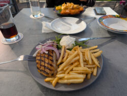 Ein Tisch im Restaurant. Auf dem Telle im Vordergrund liegt ein Bifteki mit Pommes, auf dem hinteren Teller ein Geflügelschnitzel mit Pommes. 