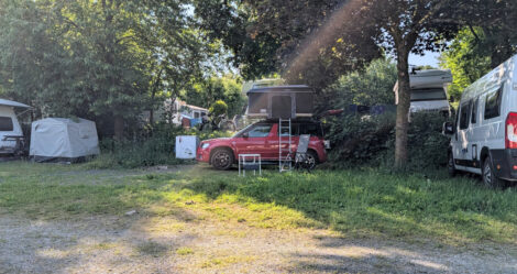 Ein PKW mit einem aufgestellten Dachzelt steht auf einem Campingplatz zwischen Wohnwagen unter einem Baum.