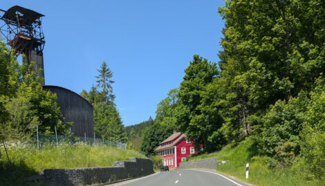 Ein alter Förderturm eines Bergwerks am Rande einer Straße im Harz. Geradeaus sieht man ein rotes Haus.