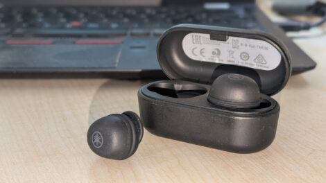 Ein Set anthrazitfahrbener In-Ear-Headsets. Ein Stöpsel steckt im offen stehenden Ladecase und ein Stöpsel liegt daneben und man kann das Yamaha-Logo erkennen. Im Hintergrund sieht man die Tastatur eines Laptops.
