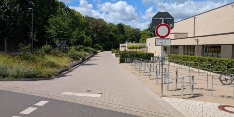 Die Zufahrt zu einem Hallen- und Freibad. Rechts sind Anschließbügel für Fahrräder zu sehen. Links ist eine mit Büschen bewachsene Böschung. Ganz unten am Bildrand sieht man die Kante zwischen Asphalt der Fahrbahn und gepflasterter Fläche vor dem Hallenbad. Ein rot-weißes Verkehrszeichen 250 verbietet die Durchfahrt.