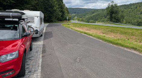 Ein roter Skoda Yeti mit einem Wohnwagen dahinter. Man sieht von vorne auf das Gespann und die Kamera fängt nur die Fahrerseite ein. Rechts sieht man die Fahrspur eines Raststreifens und rechts daneben die Straße. Im Hintergrund sind bewaldete Hügel.