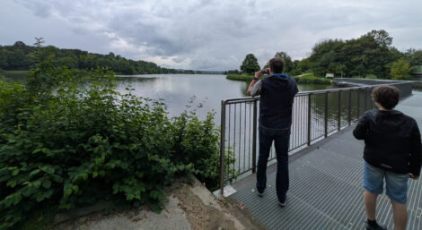 Ein Mann steht am Rand einer Metallbrücke und fotografiert einen kleinen See, der vo Bäumen umgeben ist. Rechts daneben steht einer kleiner Junge. Beide Menschen sind von hinten zu sehen.