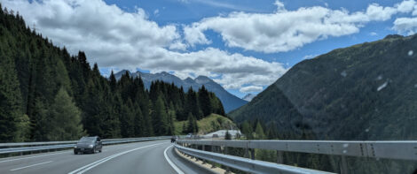 Blick von einer Autobahn auf die Schweizer Alpen mit bewaldeten Hügeln im Vordergrund unter einem blauen Himmel über den weiße Wolken ziehen.