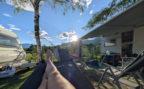 Meine Beine in einer Hängemattenliege. Direkt dahinter steht eine Birke. Rechts im Bild die Markise vor unserem Wohnwagen mit Tischen und Stühlen darunter. Die Sonne steht tief über den Bergen im Hintergrund an einem blauen Himmel.