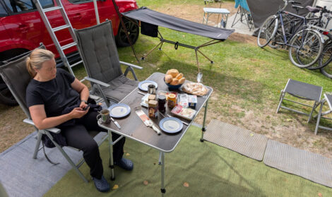 Eine Frau sitzt in einem Campingstuhl an einem Campingtisch, welcher für ein Frühstück gedeckt ist, und schaut auf ihr Smartphone.