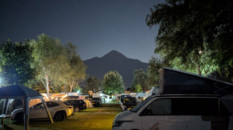 Blick in der Nacht auf einen Berg von einem Campingplatz aus. Rechts und links sieht man Büme unter denen Caravans, Zelte und Autos stehen, die von kleinen Lampen beleuchtet werden. Der Bergim Hintergrund ist als dunkler Schatten zu sehen, über dem die Sterne leuchten.