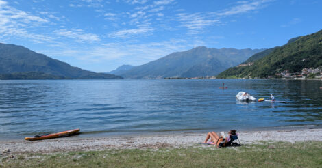 Eine Frau liegt in einem flachen Liegestuhl auf einem Rasen, der in einen Kiesstrand an einem blauen See übergeht. Am Horizont am Ende des Sees sieht man alpine Berge.