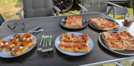Ein Campingtisch mit fünf Tellern, auf denen jeweils vier große Stücke Pizza Margarita vom Bäcker liegen.