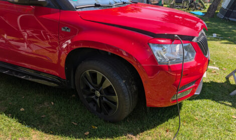 Die Front eines roten Auto, dessen Motohaube einen Spalt aufsteht. Ein Stromkabel liegt in einer Schlaufe auf dem rasen und führt dann hoch in den Motorraum.