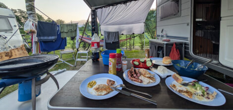 Ein Campingtisch unter einer Wohnwagenmarkise mit gegrilltem Fleich und Würstchen sowie Salat auf Tellern.