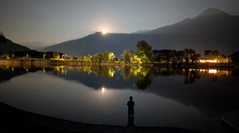 Der Mond geht über den Bergen des Comer See auf und spiegelt sich im Wasser. Bäum und Gebäude am Seeufer werden ebenfalls auf der spiegelglatten Wasseroberfläche gespiegelt. Im Vordergrund sieht man die Silhouette eines kleinen Jungen, der bis zu den Knie im Wasser steht.