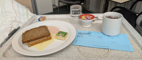 Zwei Scheiben Brot, etwas Butter, Käse und ein Joghurt auf einem Tablett in einem Krankenhauszimmer.