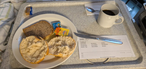 Ein Brötchen, eine Scheibe Brot, Käse, Marmelade, Yoghurt und eine Tasse Kaffee auf einem Krankenhaustablett.