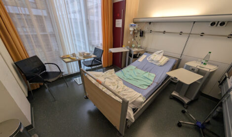 Ein Krankenbett in einem Krankenhauszimmer. Vor einem Fenster ist eine kleine Sitzecke aus zwei Stühlen und einem Tisch.