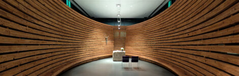 Ein halbrund aus rohen Holzwänden und einer umlaufenden Sitzbank mit einem quaderförmigen Stein mit zwei davor stehenden Stühlen bildet ein Kapelle in einem Krankenhaus.