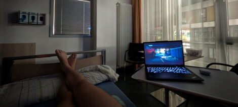 Auf dem Beistellwagen eines Krankenhausbettes steht ein aufgeklappter Laptop, auf dem eine Serie läuft. Auf dem unteren Ende des Bettes neben dem der Tisch steht, sieht man ein nacktes Bein.