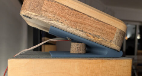 Seitenansicht eines kleinen Lautsprechers, der mit der Rückwand auf der Oberseite eines großen Standlautsprechers liegt. Damit ein ca. 20° Winkel entsteht, liegt der Lautsprecher auf einer hinten freitragenden Rampe.